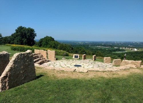 A circular stone monument surrounded by grass, overlooking a scenic valley under a clear blue sky.