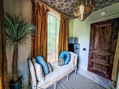Cozy corner with a bench, decorative pillows, a potted plant, and a patterned rug, featuring large windows and a chandelier.