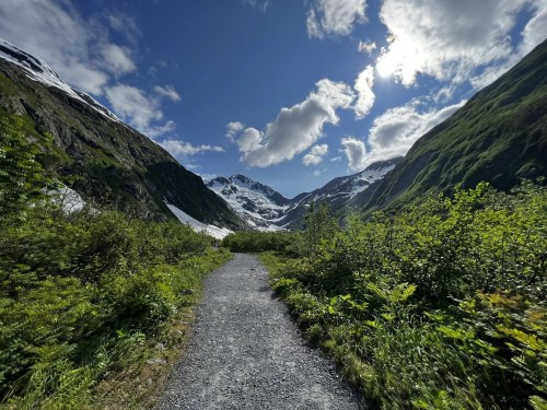 A gravel path leads through lush greenery towards snow-capped mountains under a bright blue sky with fluffy clouds.