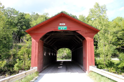 A red covered bridge with a wooden interior, surrounded by green trees and a clear blue sky.