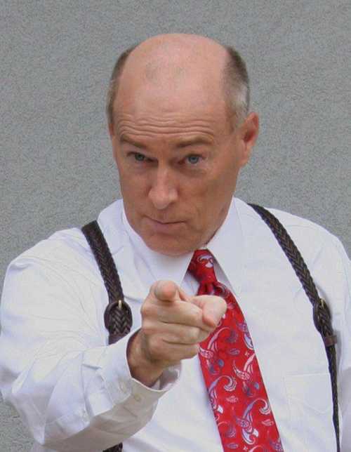 A man in a white shirt and red tie with a paisley pattern points assertively at the camera, wearing suspenders.