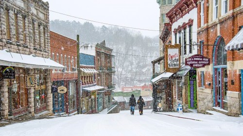 Two people walk down a snowy street lined with historic buildings and shops, creating a picturesque winter scene.