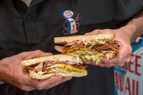 A person holds two large sandwiches filled with various meats, cheese, and lettuce, with a gas station backdrop.