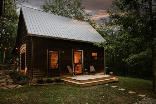 A cozy wooden cabin with a metal roof, surrounded by trees, illuminated warmly at dusk. Two chairs on the porch.