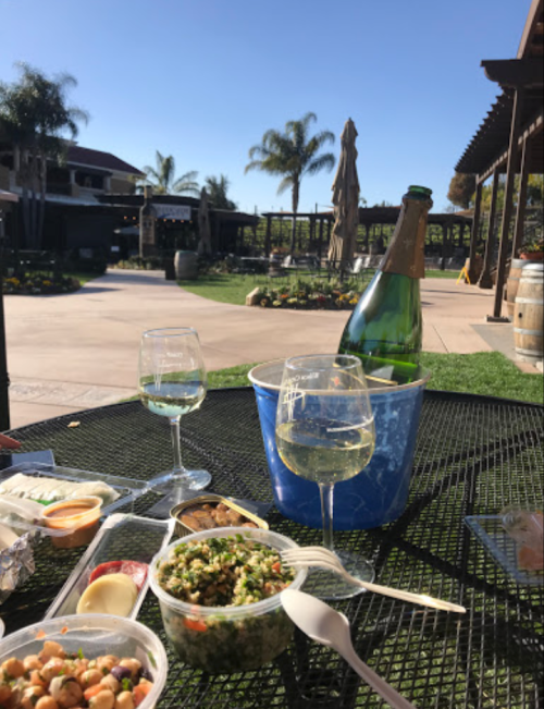 A table set outdoors with two glasses of white wine, a bottle in an ice bucket, and various food containers.