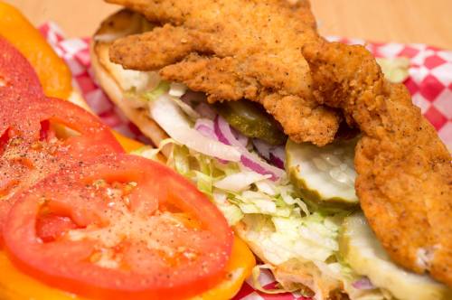 A close-up of a sandwich with fried chicken, lettuce, pickles, and sliced tomatoes on a checkered paper background.
