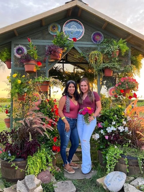 Two women stand in front of a flower-adorned gazebo, smiling and holding flowers, surrounded by vibrant greenery.