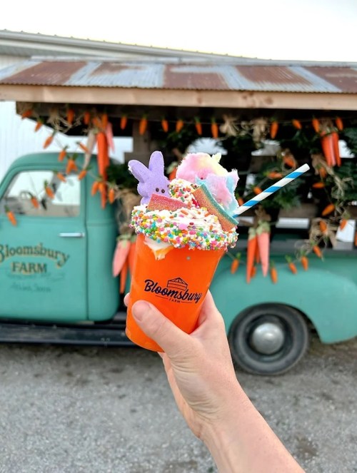 A colorful milkshake topped with whipped cream, sprinkles, and candy, held in front of a vintage truck at a farm.