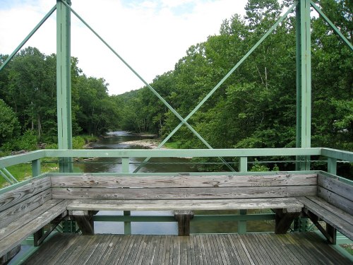 A wooden bench on a green bridge overlooking a serene river surrounded by lush trees.