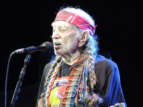 A musician with long braids and a red bandana sings into a microphone on stage.
