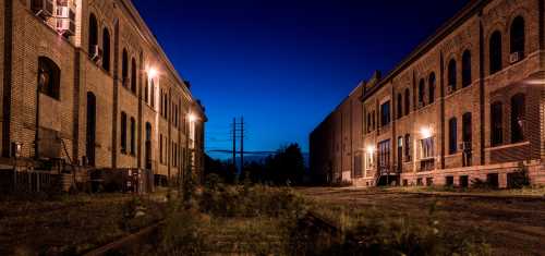 Abandoned brick buildings at dusk, illuminated by streetlights, with overgrown grass and power lines in the background.