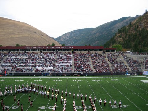 A football stadium filled with fans, with mountains in the background and a marching band performing on the field.