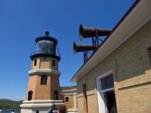 A historic lighthouse with a brick building and large horns on the roof, set against a clear blue sky.