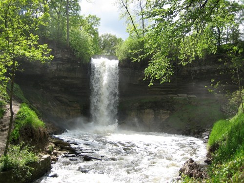 A cascading waterfall surrounded by lush green trees and rocky terrain, with mist rising from the water below.