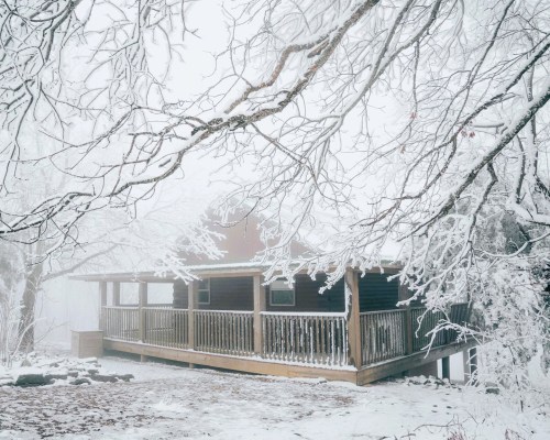 A cozy wooden cabin surrounded by snow-covered trees in a foggy winter landscape.