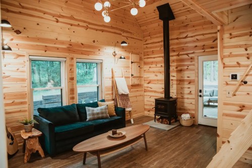 Cozy wooden cabin interior featuring a green sofa, round coffee table, and a wood stove, surrounded by large windows.