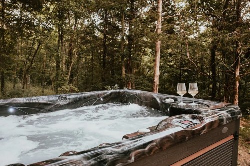 A hot tub surrounded by trees, with two glasses on the edge, creating a serene outdoor relaxation space.