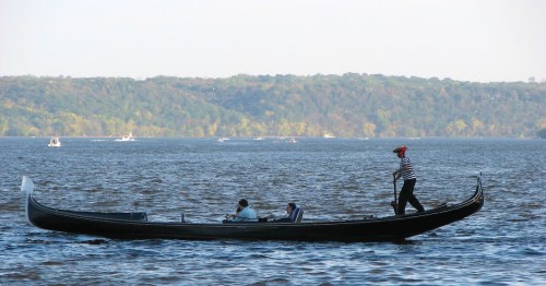 A gondola on a calm river with a person in striped attire rowing, surrounded by trees in autumn colors.