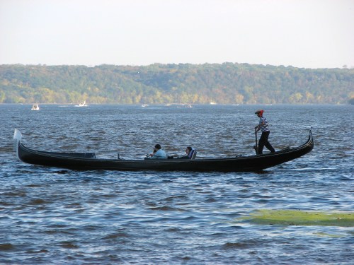 A gondola on a calm lake with two passengers and a gondolier in a striped shirt, surrounded by trees in the background.