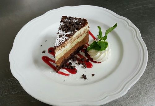 A slice of layered chocolate cake on a white plate, garnished with mint, whipped cream, and raspberry sauce.