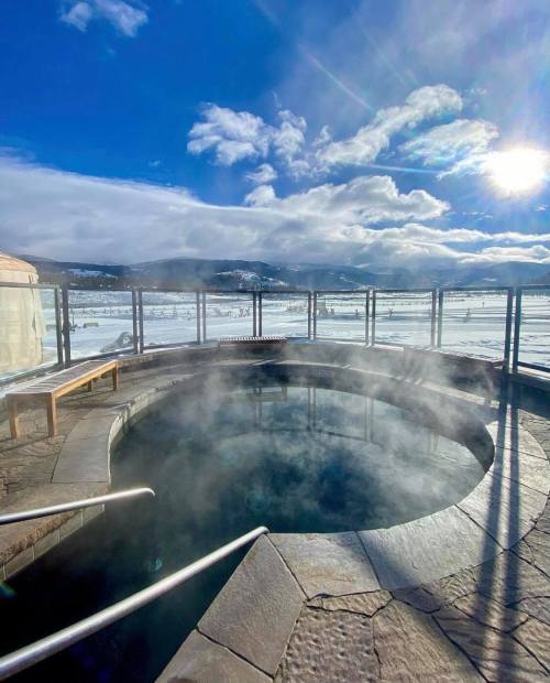 A steaming hot tub on a snowy landscape, with mountains and a bright blue sky in the background.