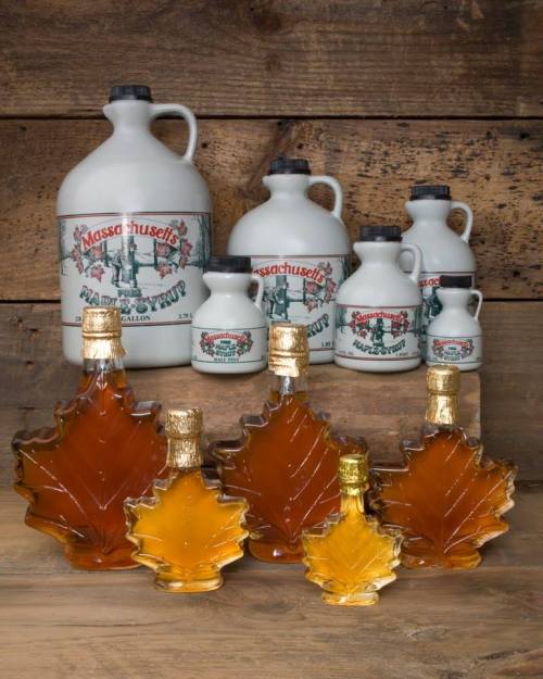 A collection of maple syrup bottles in various sizes, including glass leaf-shaped bottles and ceramic jugs, on a wooden background.