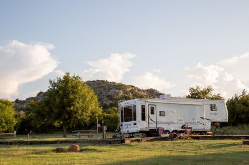 A white RV parked in a grassy area with trees and a rocky hill in the background under a blue sky with clouds.