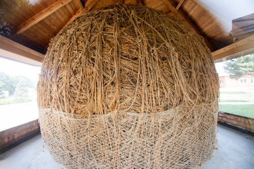 A large, spherical sculpture made of intertwined natural fibers, displayed in a wooden structure.