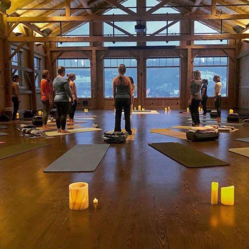 A group of people practicing yoga in a wooden studio, surrounded by candles and mats, with large windows showing a scenic view.