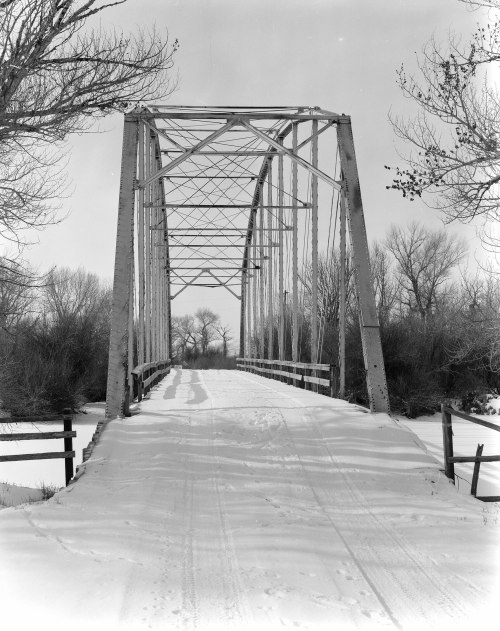A snow-covered bridge with a metal frame spans a frozen river, surrounded by bare trees in a winter landscape.
