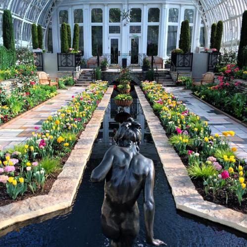 A serene garden with colorful flowers, a statue in a pond, and a glass conservatory in the background.