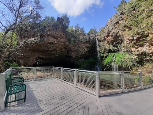 A wooden deck with green benches overlooks a serene waterfall and lush greenery in a rocky landscape.