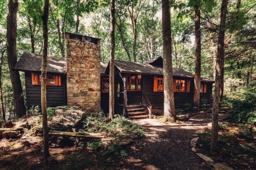 A rustic cabin with a stone chimney, surrounded by lush green trees and a winding path in a forested area.