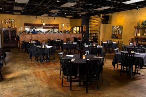 A spacious restaurant with black tables and chairs, warm yellow walls, and a bar area in the background.