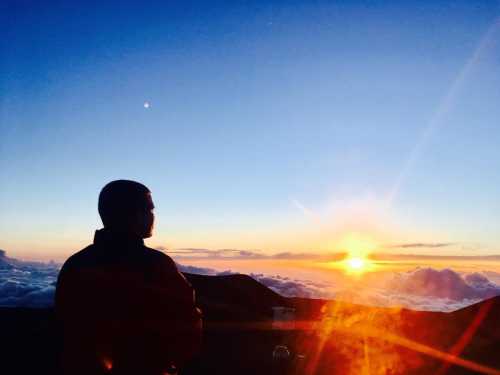 A person silhouetted against a vibrant sunrise above the clouds, with a clear sky and distant stars visible.