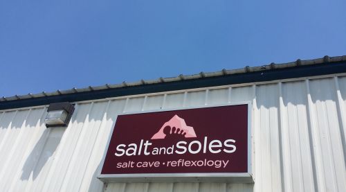 Sign for "Salt and Soles" featuring a mountain graphic, promoting salt cave and reflexology services against a blue sky.