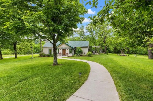 A charming house with a green metal roof, surrounded by lush trees and a manicured lawn, with a winding pathway leading to it.