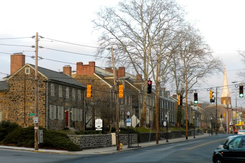 Historic stone buildings line a quiet street, with traffic lights and a church steeple visible in the background.