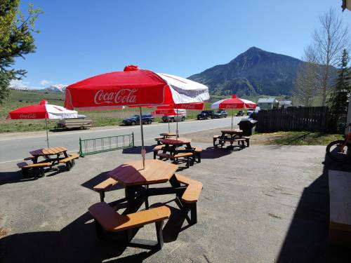 Outdoor seating area with picnic tables and red umbrellas, set against a backdrop of mountains and clear blue sky.