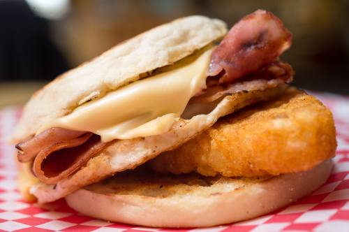 A close-up of a breakfast sandwich with a crispy patty, ham, cheese, and an English muffin on a red checkered plate.