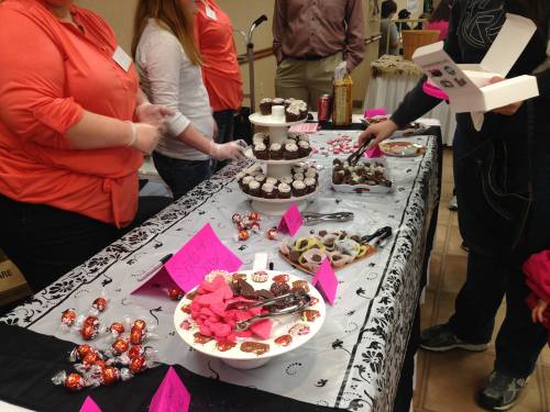 A dessert table featuring cupcakes, candies, and chocolates, with people sampling treats and colorful labels.