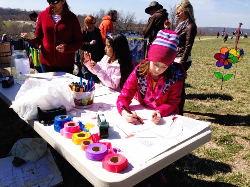 Children are crafting at a table outdoors, surrounded by adults and colorful decorations in a grassy field.