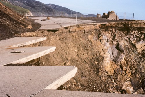 A damaged road with eroded edges, revealing rocky terrain and a mountainous landscape in the background.
