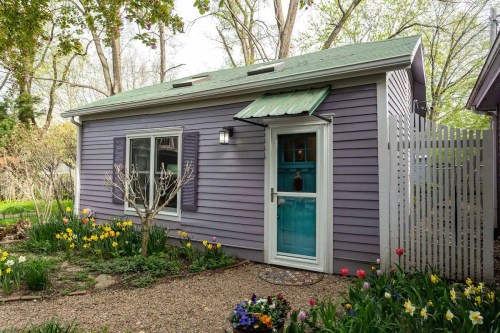 A charming purple cottage with a green roof, surrounded by blooming flowers and a white picket fence.