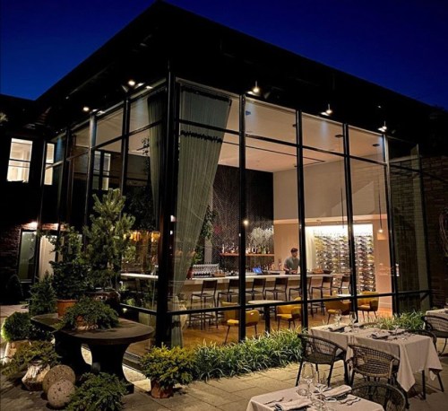 A modern restaurant with large glass windows, dim lighting, and outdoor seating, set against a night sky.