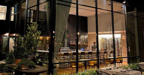A modern restaurant with glass walls, featuring a bar, seating, and greenery outside, illuminated at night.