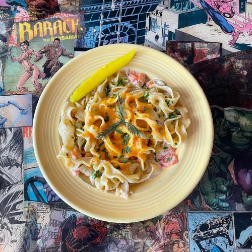 A plate of creamy pasta topped with herbs and a lemon slice, set on a colorful comic book background.