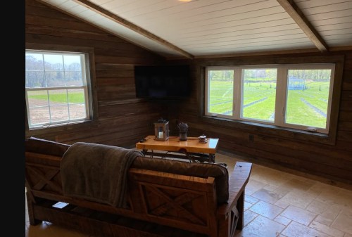 Cozy interior of a rustic room with a couch, table, and large windows overlooking a green field.