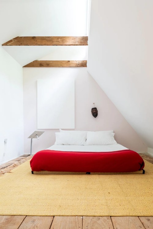 A minimalist bedroom with a red bed, wooden beams, and a large area rug, featuring a simple wall art piece.