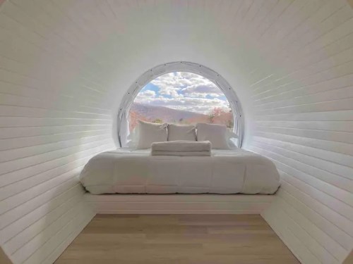 Cozy, minimalist bedroom with a circular window showcasing a scenic view of mountains and clouds.